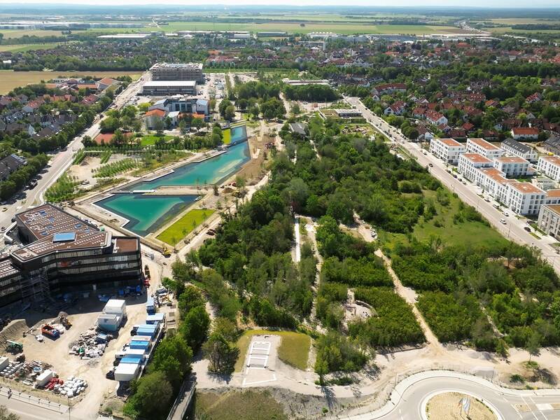 Luftbild: Ein See führt in mitten einer Wiese durch. Links und rechts daneben befindet sich eine Baustelle.