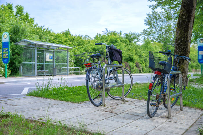 Zwei Fahrräder sind im Vordergrund angeschlossen. Im Hintergrund erkennt man eine Bushaltestelle.